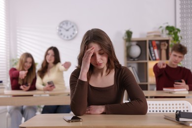 Photo of Cyber bullying. Upset teenage girl with smartphone and schoolmates pointing at her in classroom, selective focus