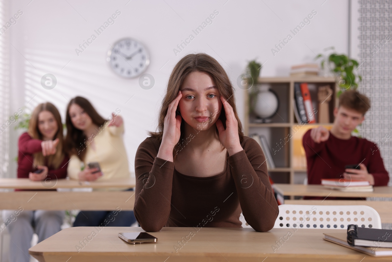 Cyber bullying. Upset teenage girl with smartphone and schoolmates pointing at her in classroom, selective focus Photo of Cyber bullying. Upset teenage girl with smartphone and schoolmates pointing at her in classroom, selective focus