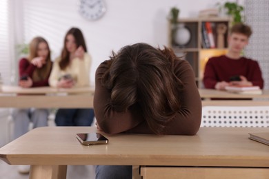 Photo of Cyber bullying. Upset teenage girl with smartphone and schoolmates pointing at her in classroom, selective focus