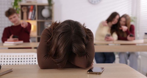 Photo of Cyber bullying. Upset teenage girl with smartphone and schoolmates pointing at her in classroom, selective focus