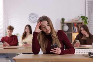 Photo of Cyber bullying. Upset teenage girl with smartphone at desk in classroom