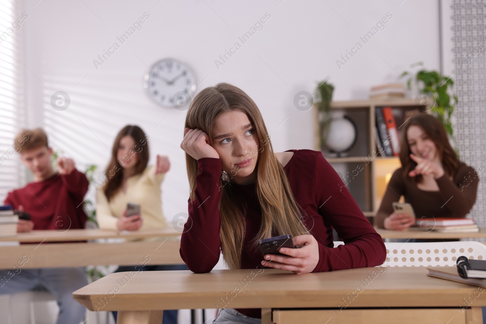 Cyber bullying. Upset teenage girl with smartphone and schoolmates pointing at her in classroom, selective focus Photo of Cyber bullying. Upset teenage girl with smartphone and schoolmates pointing at her in classroom, selective focus
