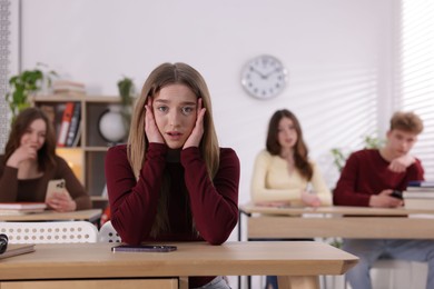 Photo of Cyber bullying. Upset teenage girl with smartphone at desk in classroom