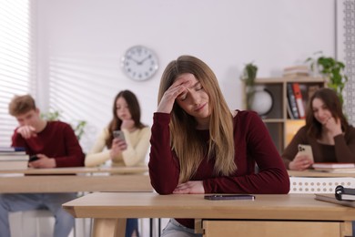 Photo of Cyber bullying. Upset teenage girl with smartphone at desk in classroom