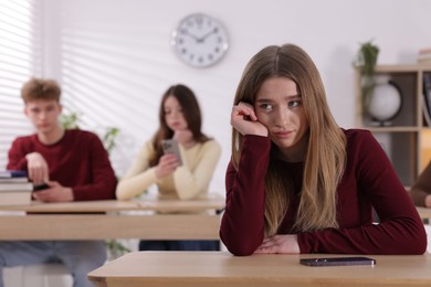 Photo of Cyber bullying. Upset teenage girl with smartphone at desk in classroom