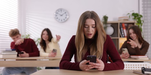 Photo of Cyber bullying. Upset teenage girl with smartphone and schoolmates pointing at her in classroom, selective focus