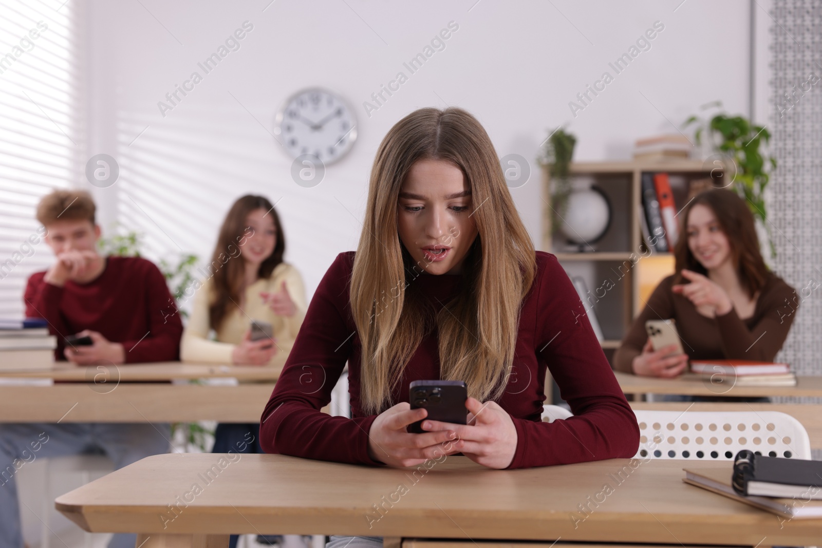 Cyber bullying. Upset teenage girl with smartphone and schoolmates pointing at her in classroom, selective focus Photo of Cyber bullying. Upset teenage girl with smartphone and schoolmates pointing at her in classroom, selective focus