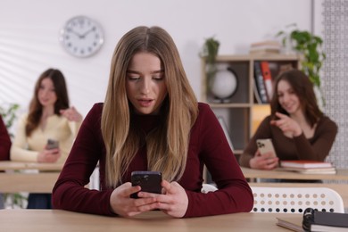 Photo of Cyber bullying. Upset teenage girl with smartphone and schoolmates pointing at her in classroom, selective focus