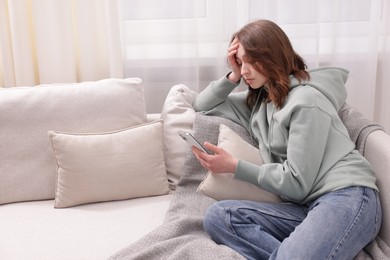 Photo of Cyber bullying. Upset teenage girl with smartphone on sofa at home. Space for text