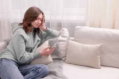 Photo of Cyber bullying. Upset teenage girl with smartphone on sofa at home. Space for text