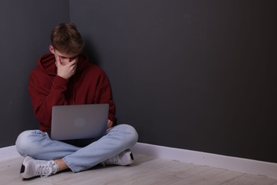 Photo of Cyber bullying. Upset teenage boy with laptop on floor indoors. Space for text