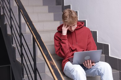Photo of Cyber bullying. Upset teenage boy with laptop on stairs indoors