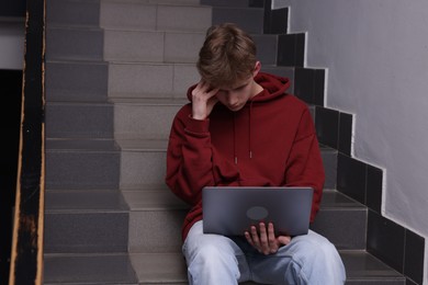 Photo of Cyber bullying. Upset teenage boy with laptop on stairs indoors