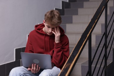 Photo of Cyber bullying. Upset teenage boy with laptop on stairs indoors