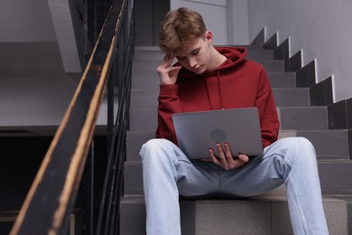 Photo of Cyber bullying. Upset teenage boy with laptop on stairs indoors