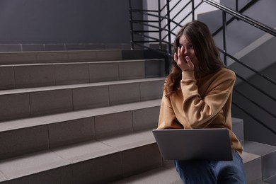 Photo of Cyber bullying. Upset teenage girl with laptop on stairs indoors. Space for text