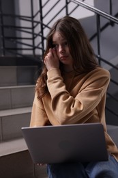 Photo of Cyber bullying. Upset teenage girl with laptop on stairs indoors