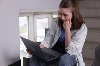 Photo of Cyber bullying. Upset teenage girl with laptop on stairs indoors. Space for text
