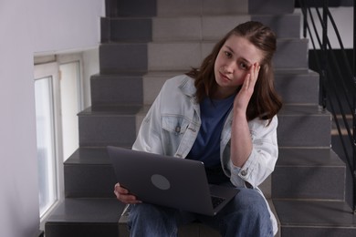 Photo of Cyber bullying. Upset teenage girl with laptop on stairs indoors. Space for text
