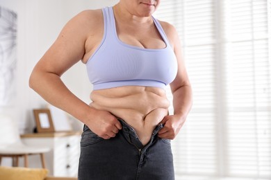 Photo of Overweight woman trying to put on tight jeans at home, closeup
