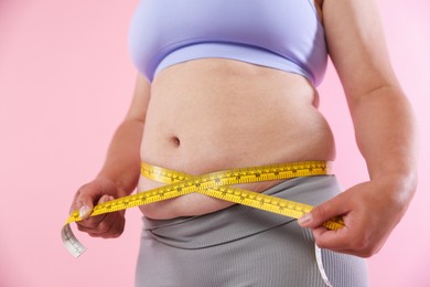 Photo of Overweight woman measuring her belly with tape on pink background, closeup