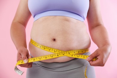 Photo of Overweight woman measuring her belly with tape on pink background, closeup
