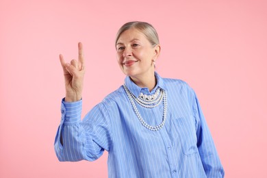 Photo of Senior woman wearing elegant pearl jewelry and making rock gesture on pink background