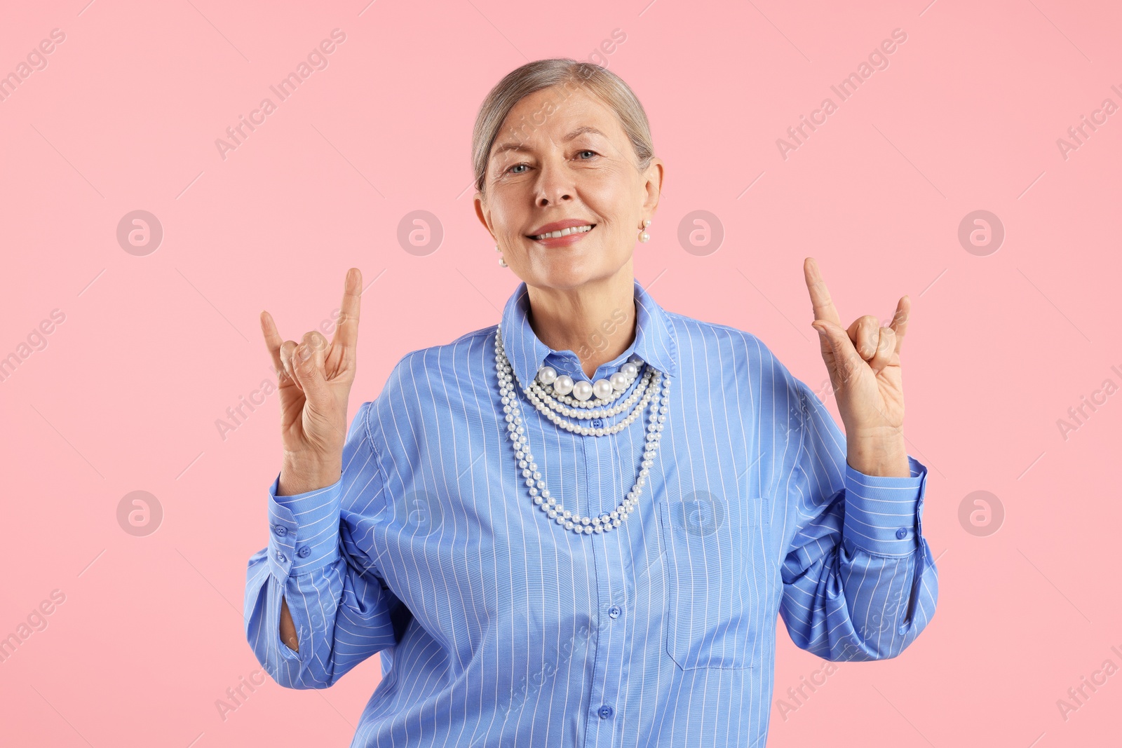 Senior woman wearing elegant pearl jewelry and making rock gesture on pink background Photo of Senior woman wearing elegant pearl jewelry and making rock gesture on pink background