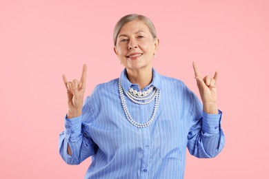Photo of Senior woman wearing elegant pearl jewelry and making rock gesture on pink background