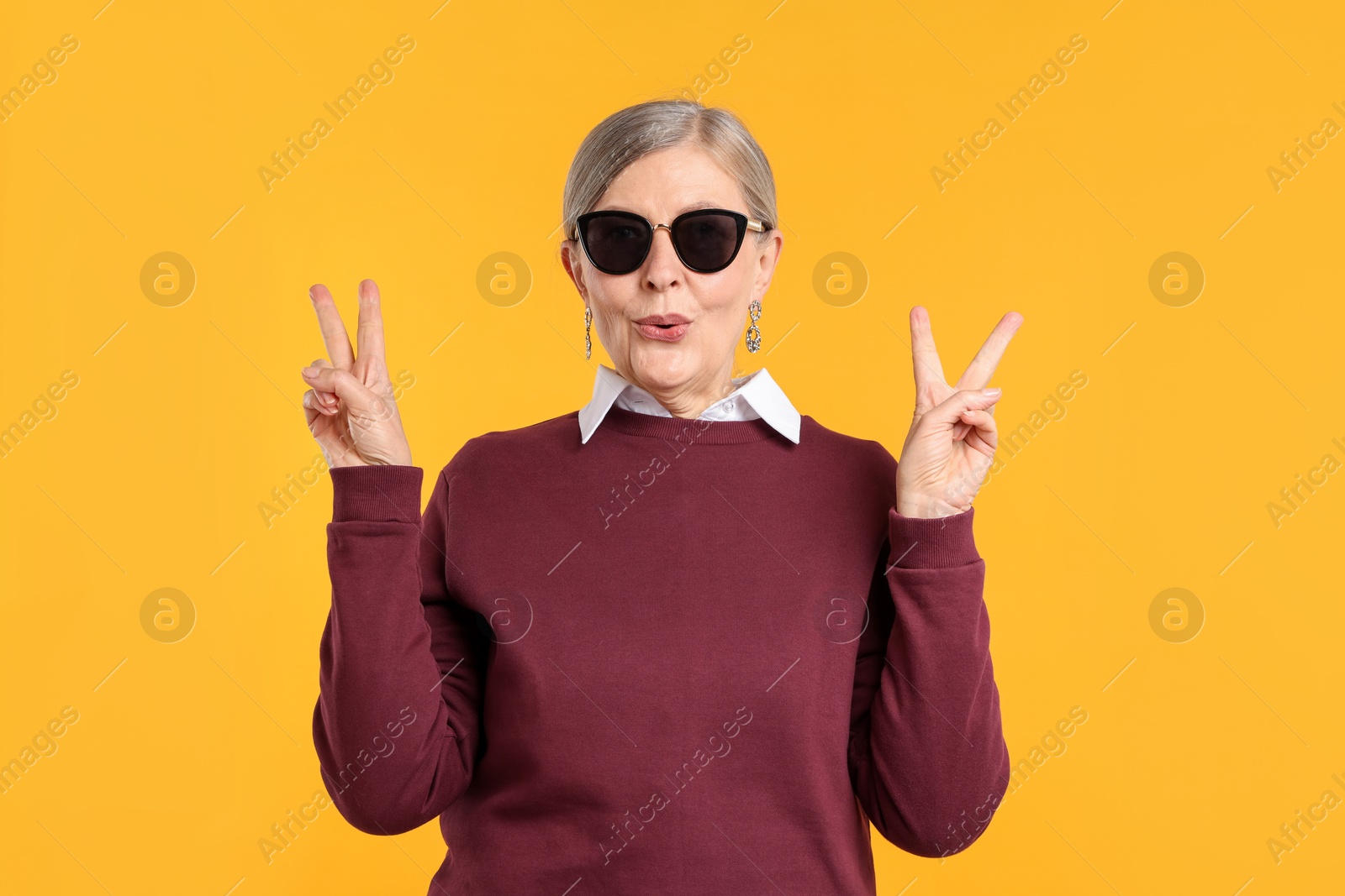 Senior woman in stylish outfit showing V-signs on orange background Photo of Senior woman in stylish outfit showing V-signs on orange background
