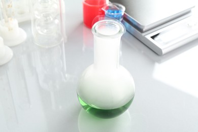 Photo of Flask with green liquid, other laboratory glassware and scale on white mirror table, closeup
