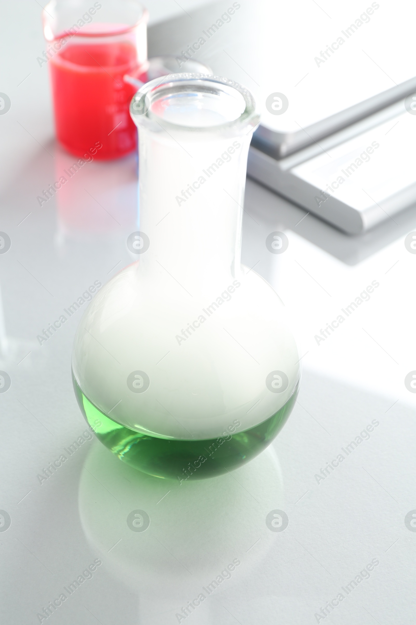 Flask with green liquid, other laboratory glassware and scale on white mirror table, closeup Photo of Flask with green liquid, other laboratory glassware and scale on white mirror table, closeup