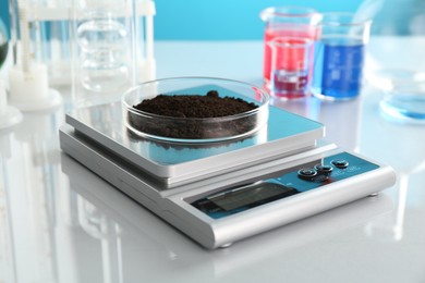 Photo of Scale with Petri dish of sample and other laboratory glassware on white mirror table against light blue background, closeup
