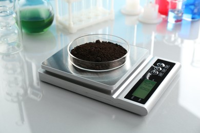 Photo of Scale with Petri dish of sample and other laboratory glassware on white mirror table, closeup