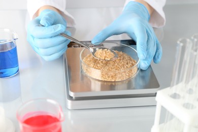 Photo of Scientist weighing Petri dish with sample on scales at white mirror table, closeup