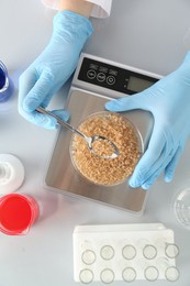 Photo of Scientist weighing Petri dish with sample on scales at white table, top view