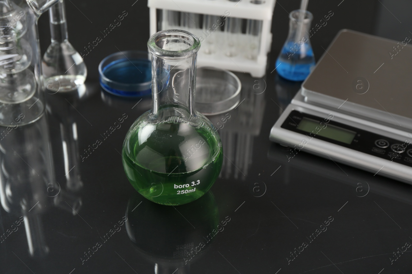 Flask with green liquid, other laboratory glassware and scale on black mirror table, closeup Photo of Flask with green liquid, other laboratory glassware and scale on black mirror table, closeup
