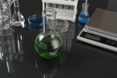 Photo of Flask with green liquid, other laboratory glassware and scale on black mirror table, closeup