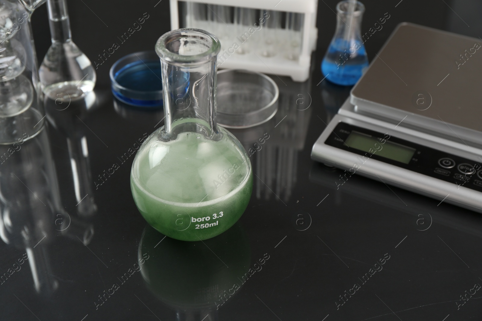Flask with green liquid, other laboratory glassware and scale on black mirror table, closeup Photo of Flask with green liquid, other laboratory glassware and scale on black mirror table, closeup