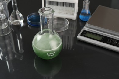 Photo of Flask with green liquid, other laboratory glassware and scale on black mirror table, closeup