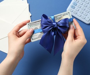 Photo of Money bonus. Woman holding dollar banknotes with bow on blue background, closeup
