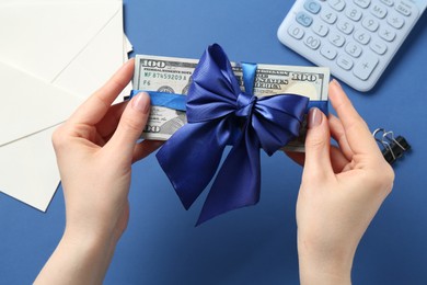 Photo of Money bonus. Woman holding dollar banknotes with bow on blue background, closeup