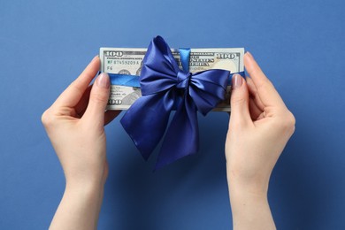 Photo of Money bonus. Woman holding dollar banknotes with bow on blue background, top view