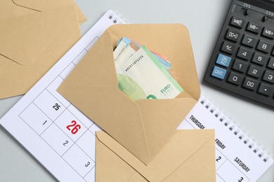 Photo of Money bonus. Envelopes with euro banknotes, calculator and calendar on light grey background, flat lay