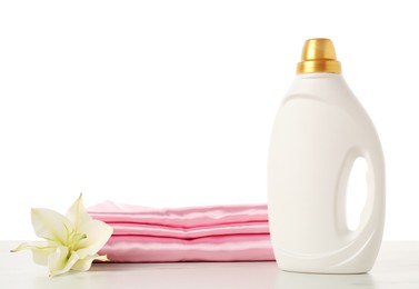 Photo of Bottle of fabric softener, stacked clothes and flower on marble table against white background