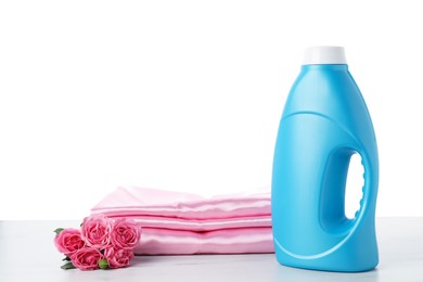 Photo of Bottle of fabric softener, stacked clothes and flowers on marble table against white background