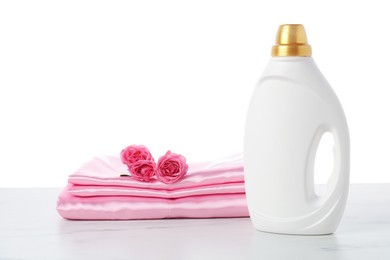 Photo of Bottle of fabric softener, stacked clothes and flowers on marble table against white background