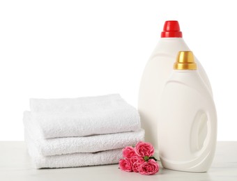 Photo of Bottles of fabric softener, stacked towels and flowers on marble table against white background