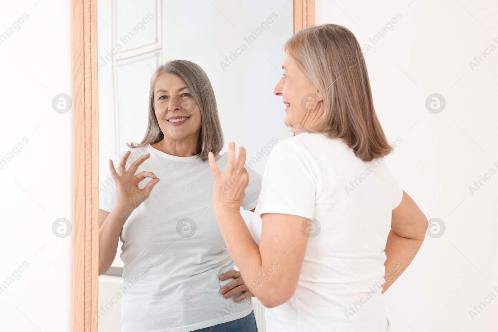 Beautiful senior woman showing ok gesture near mirror indoors Photo of Beautiful senior woman showing ok gesture near mirror indoors