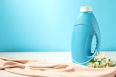 Photo of Bottle of fabric softener, clean cloth and lily flowers on white wooden table against light blue background. Space for text
